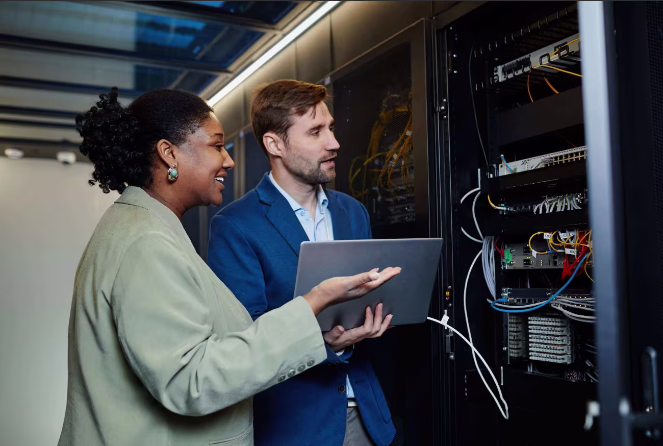 A business consultant shows a laptop to a smiling colleague beside an open server rack, illustrating improved network operations