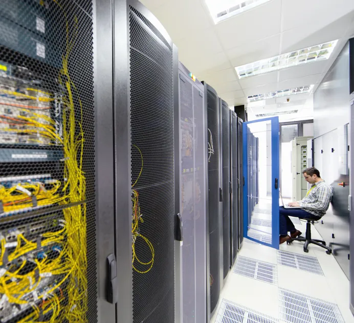 Server racks line a data centre corridor, with a technician on a laptop, representing its network consulting services in practice